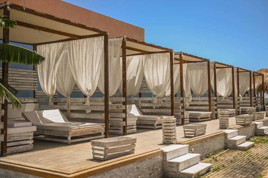 Outdoor wooden lounge chairs with white canopies, arranged in rows on a paved area with stone steps leading up to a building.