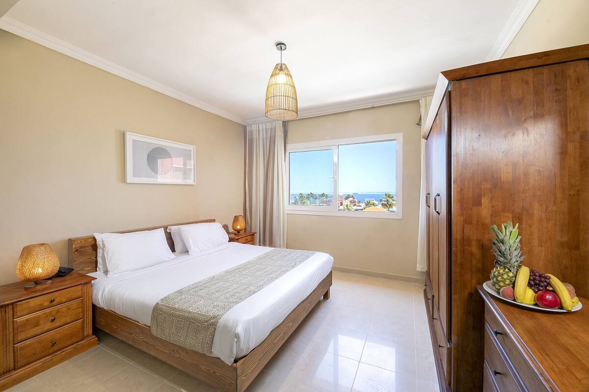 Une chambre à coucher avec un lit, des lampes de chevet, une armoire, une assiette de fruits, une fenêtre donnant sur un beau paysage et un luminaire au plafond.