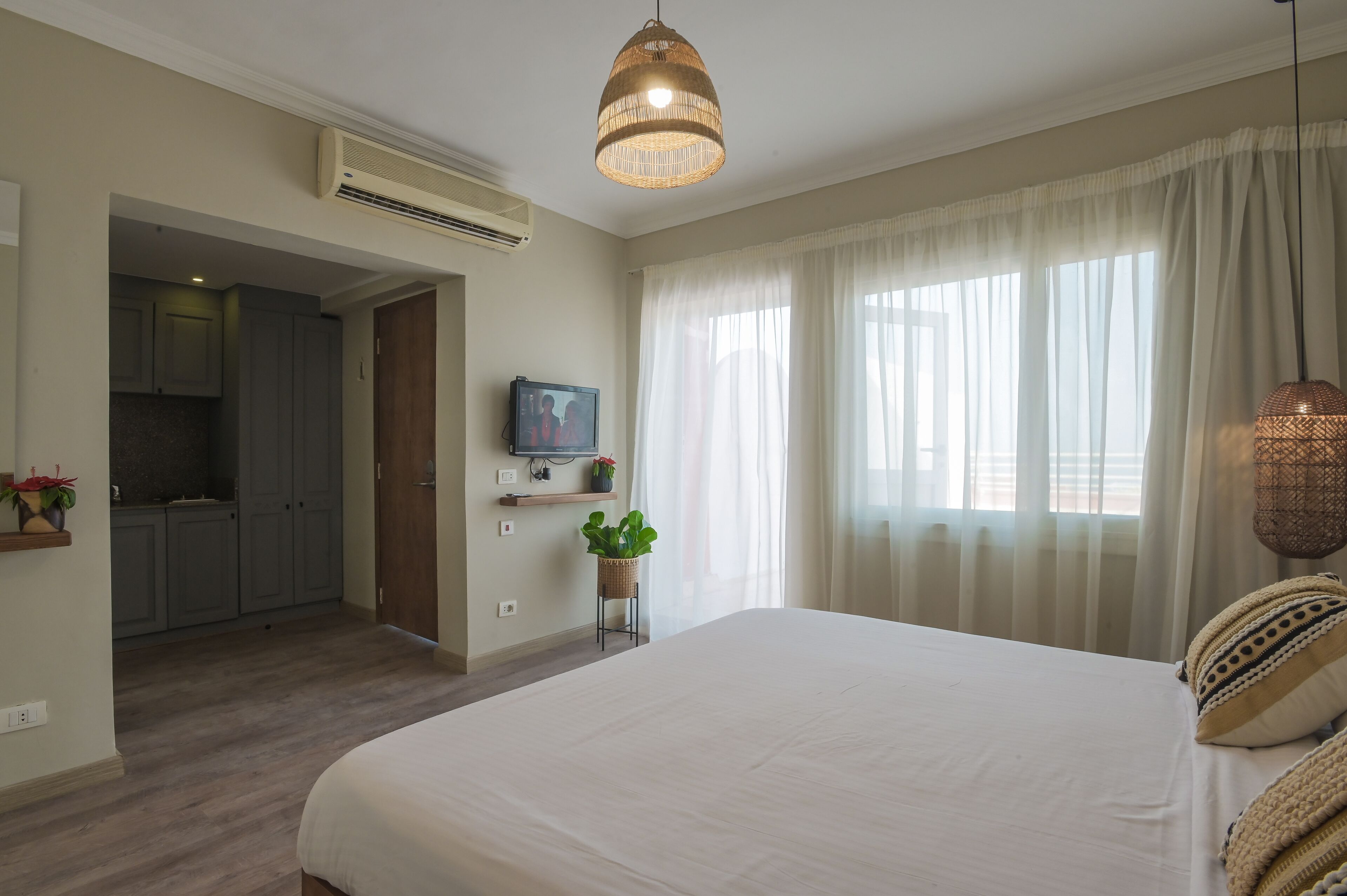 A bedroom with a large bed, a television on a stand, a hanging light fixture, and a view of a kitchen and bathroom through an open door.
