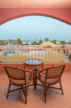 A balcony with wicker chairs and a glass table, overlooking a marina with boats and palm trees.