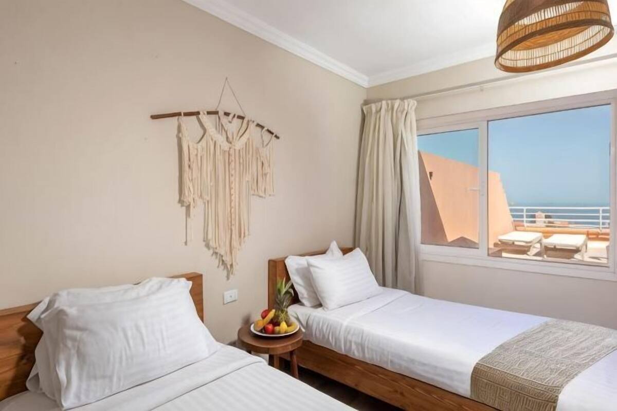 Une chambre d’hôtel avec deux lits, une petite table avec des fruits, une décoration murale en macramé et une vue sur la mer.