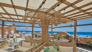 Un espace de restauration en plein air, doté de mobilier en osier et de lampes suspendues, offrant une vue sur une plage et l’océan.