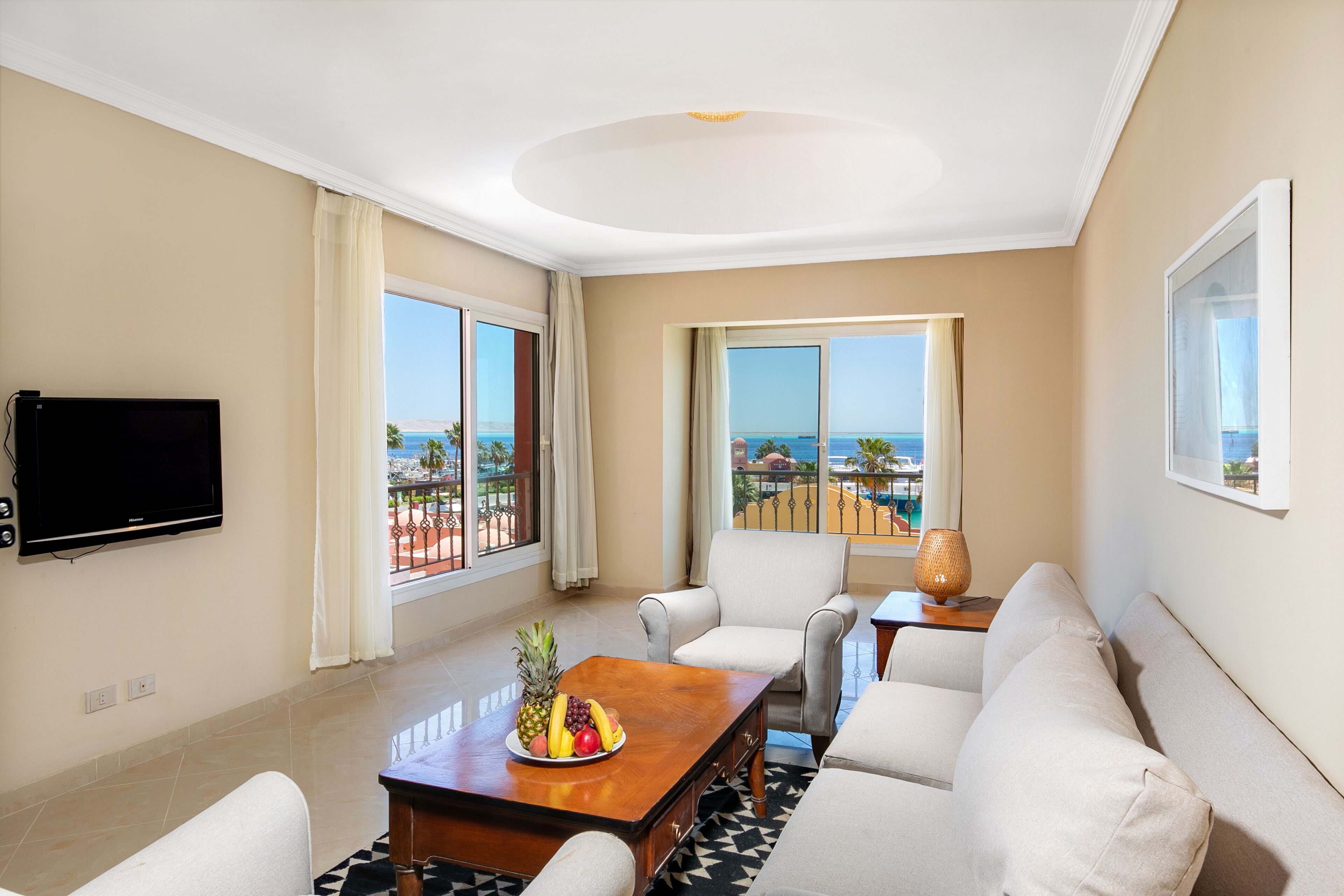 A spacious living room with a large flat-screen TV, a wooden coffee table with fruit, a sofa, and a view of the ocean through large windows.