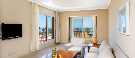A spacious living room with a large flat-screen TV, a wooden coffee table with fruit, a sofa, and a view of the ocean through large windows.
