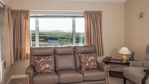 A living room with a brown leather sofa, two patterned cushions, a side table with a lamp, and a view of green fields through a large window. - 3 bedroom accommodation in Aberdaron (Aberdaron)