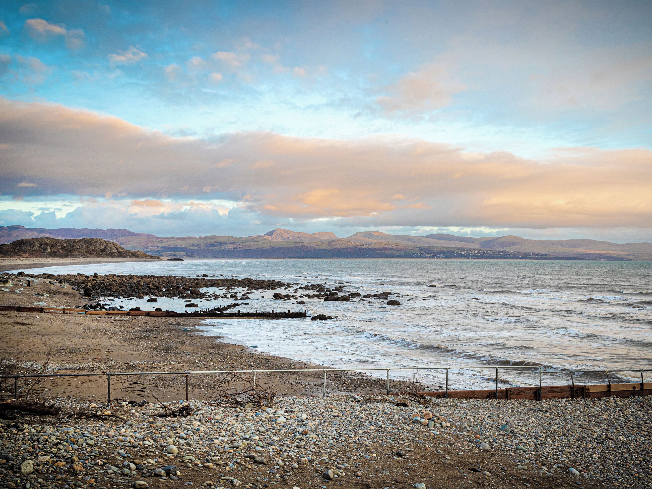 Une plage rocheuse bordée d’une clôture en bois, une mer calme et des montagnes lointaines sous un ciel nuageux.