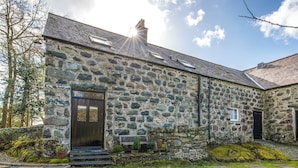 A stone building with a wooden door, steps leading to the entrance, and a garden with stepping stones. - Bwthyn Stabal-QC1199 (Criccieth)