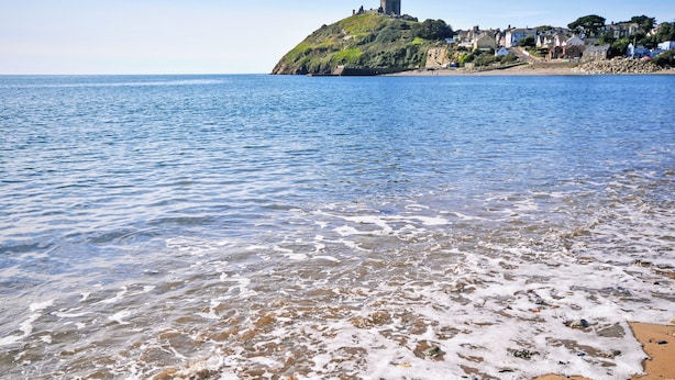 A beach with clear water, a sandy shore, and a hill with buildings in the background. - Bwthyn Stabal-QC1199 (Criccieth)