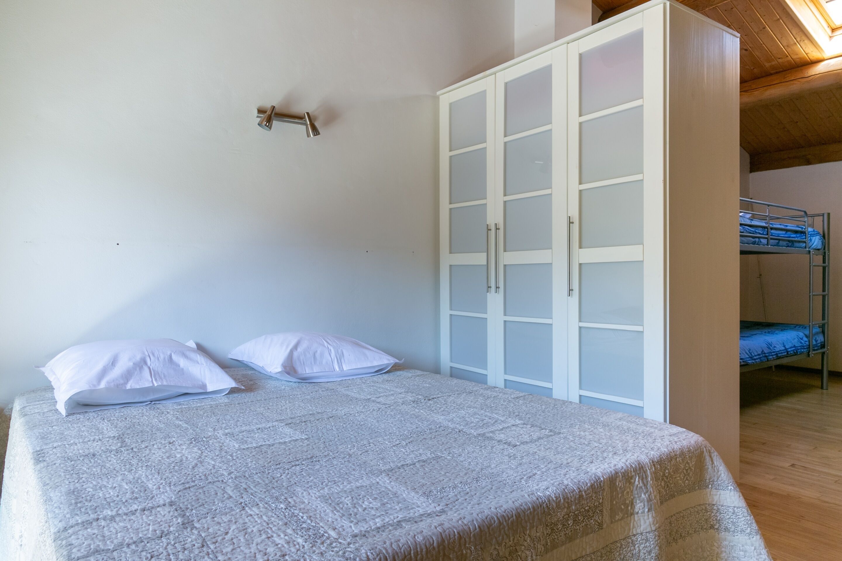 A bedroom with a bed, two pillows, a wardrobe, and a bunk bed in the background.