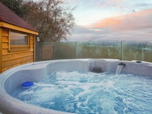 Ein Whirlpool mit Holzterrasse und Glaseinfriedung, umgeben von einem Zaun und Bäumen.