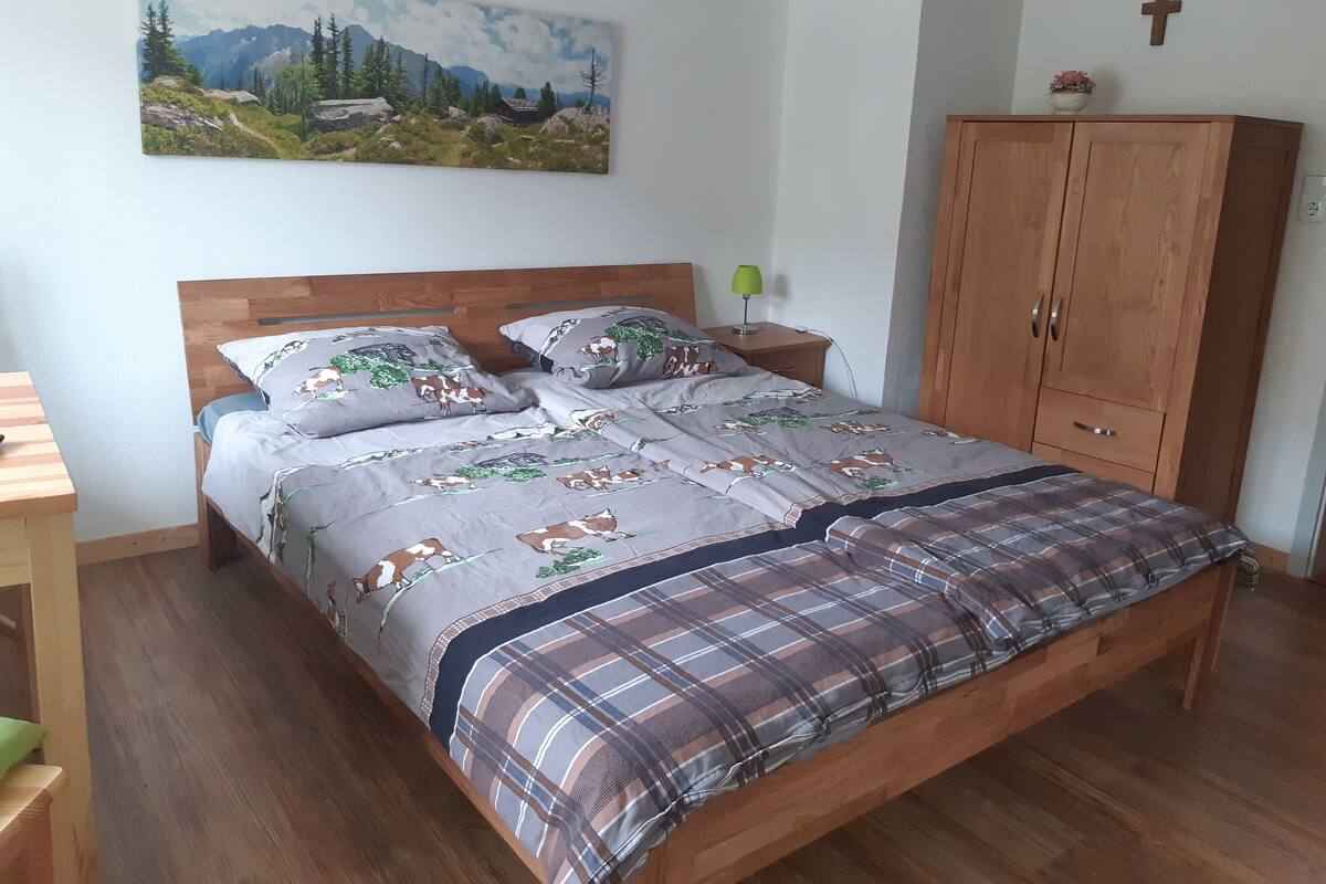 A bedroom with a wooden bed, a painting of a mountain landscape, a wooden wardrobe, and a cross on the wall.