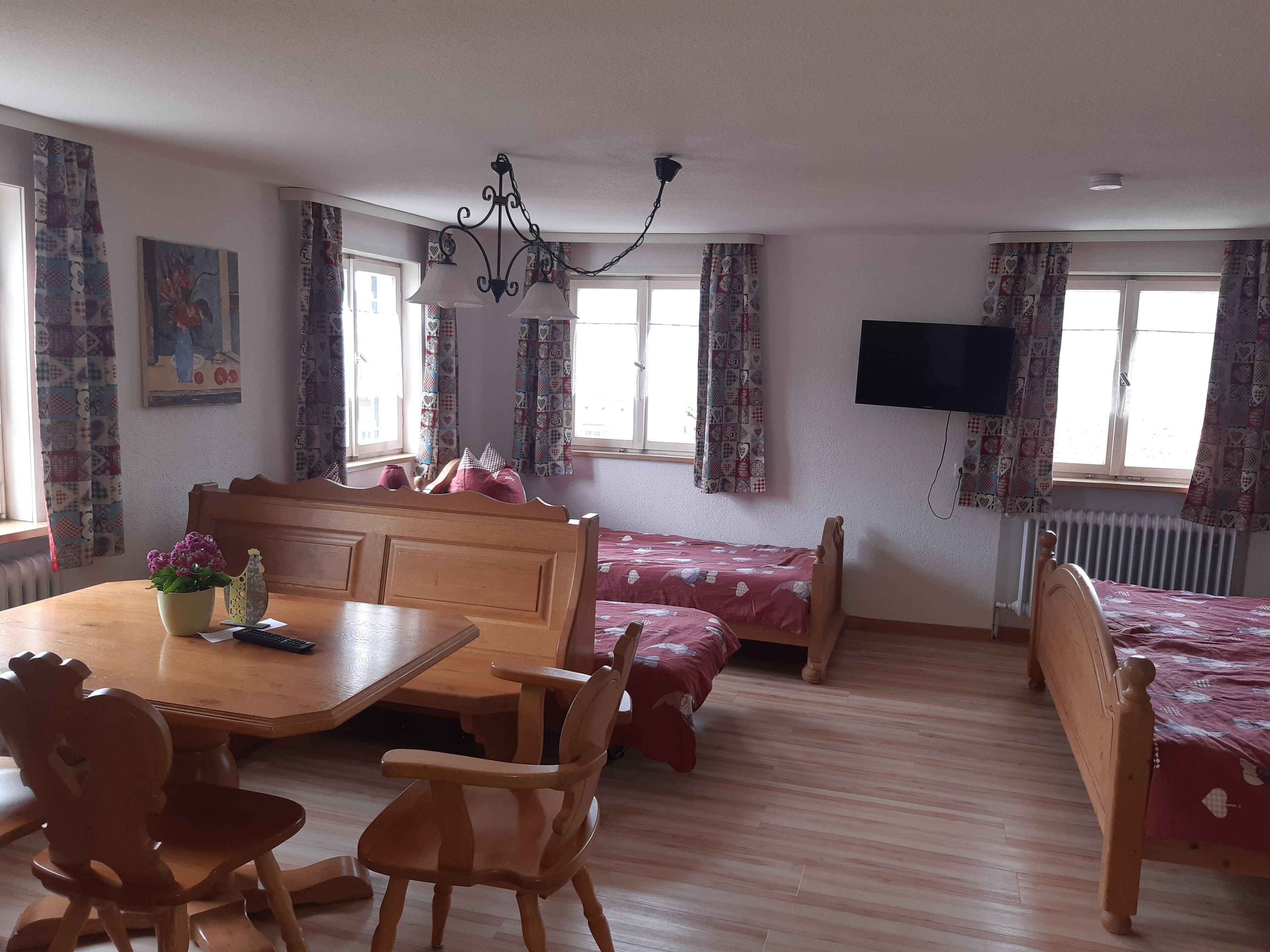 Een kamer met een houten eettafel, stoelen, een bed met een rode sprei, een aan de muur bevestigde televisie en gordijnen met een patroon.