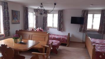 A room with a wooden dining table, chairs, a bed with a red bedspread, a TV mounted on the wall, and patterned curtains.