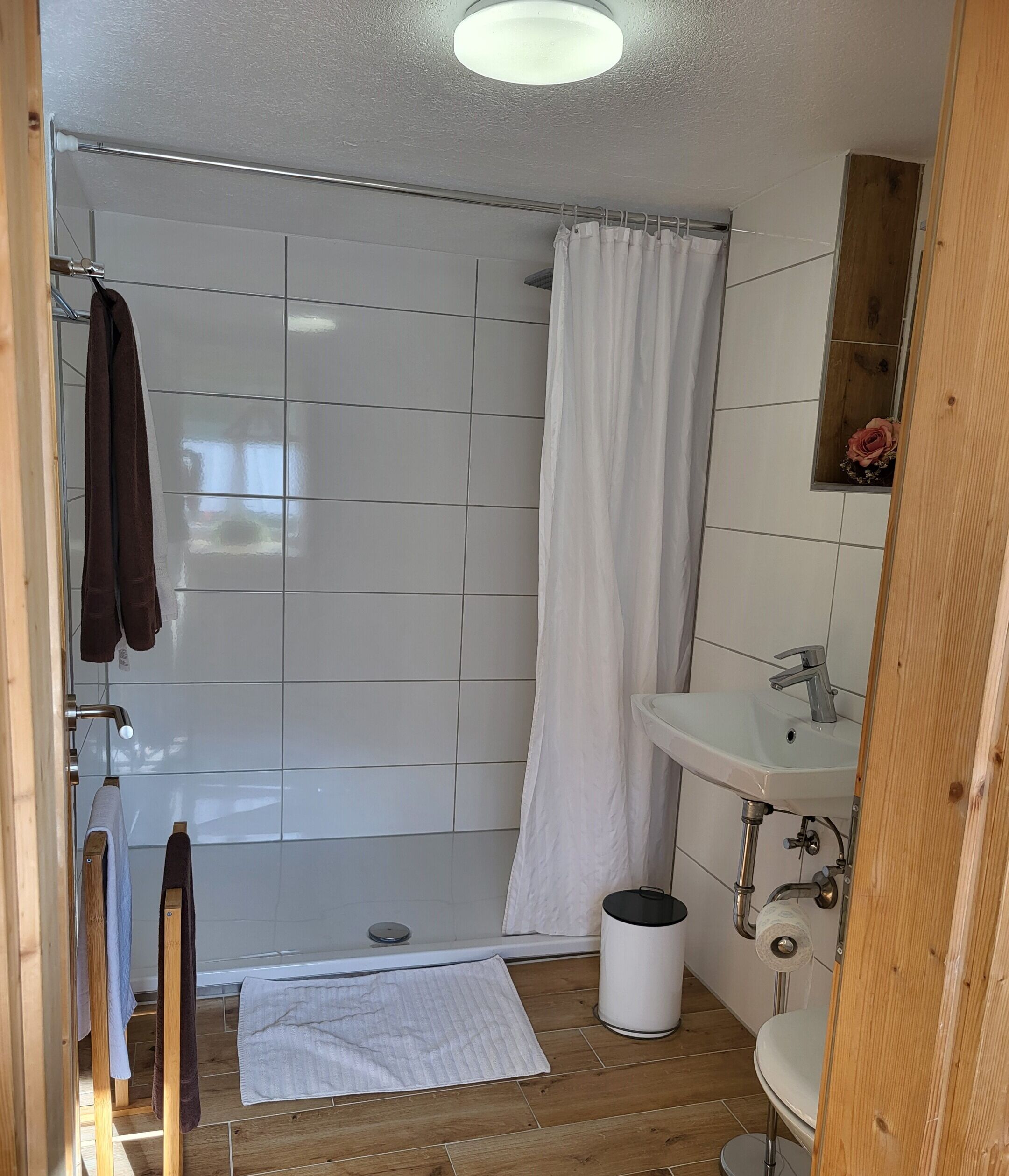 Ein Badezimmer mit Dusche, Waschbecken und Toilette. Handtücher hängen an einem Ablagegestell und ein Mülleimer steht auf dem Boden.