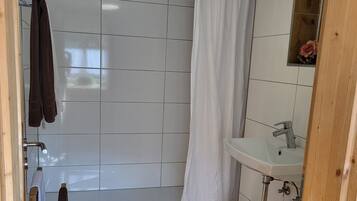 Ein Badezimmer mit Dusche, Waschbecken und Toilette. Handtücher hängen an einem Ablagegestell und ein Mülleimer steht auf dem Boden.