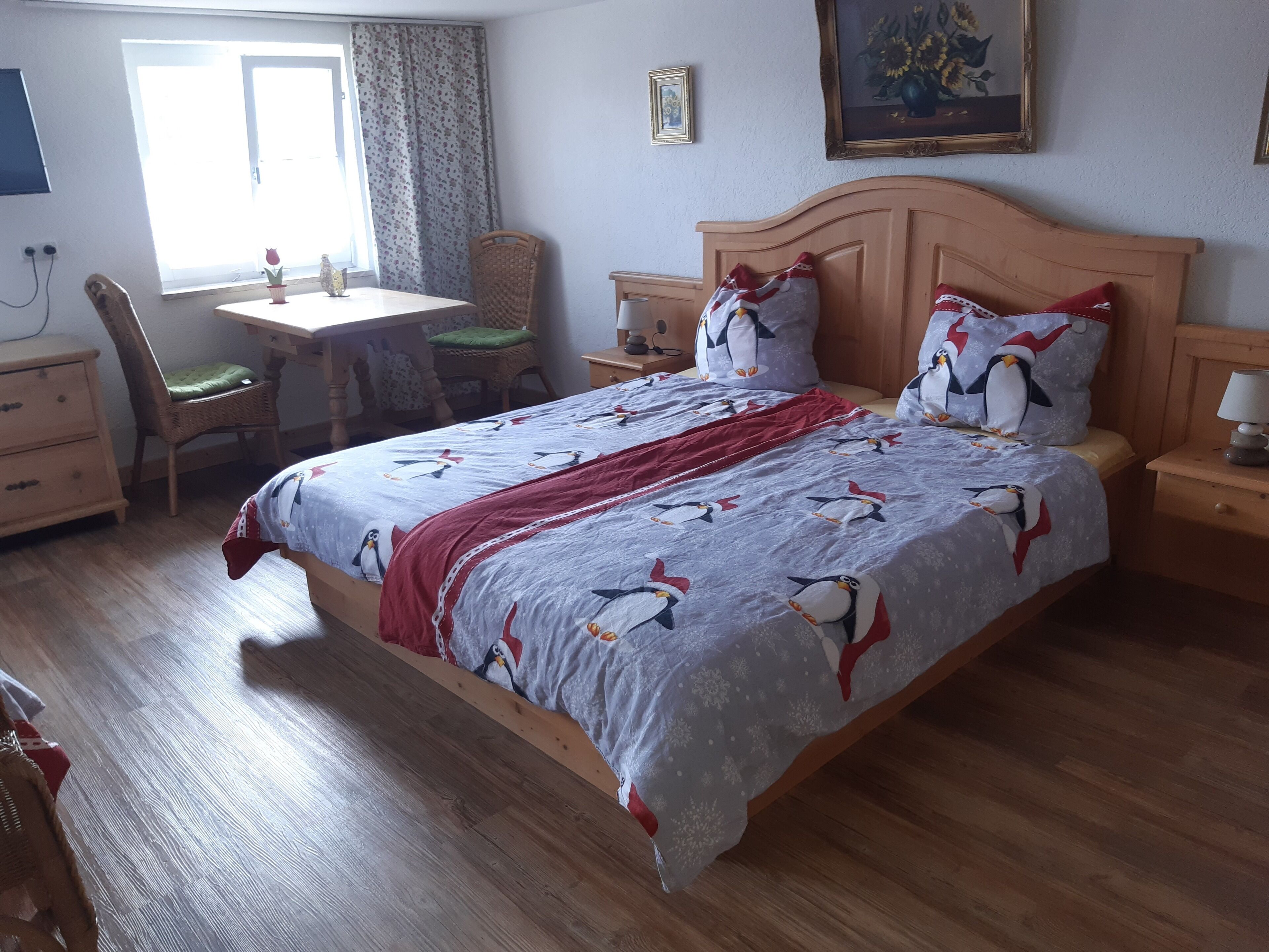 A neatly arranged bedroom with a large bed, a wooden nightstand, a bedside table, a chair, a small table, and a window with curtains.