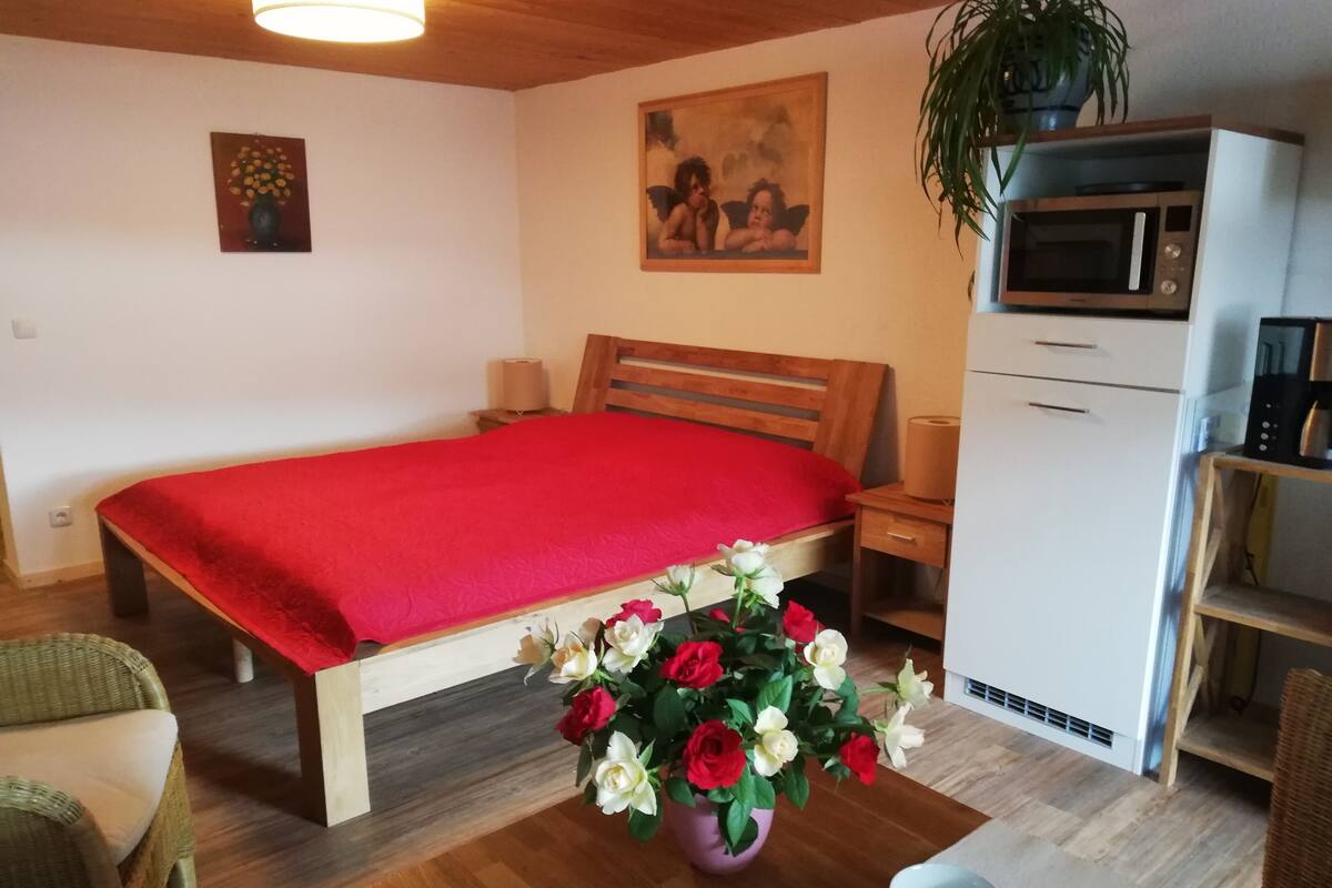 A bedroom with a wooden bed, a red bedspread, a nightstand, a bedside table, a microwave, a refrigerator, a potted plant, a vase with flowers, and a wicker chair.
