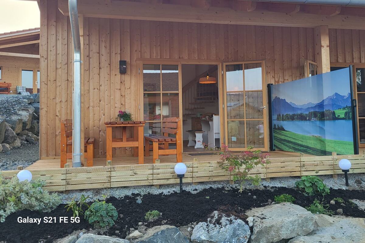 A wooden cabin with a deck, a dining area, and a large window with a mountain view.