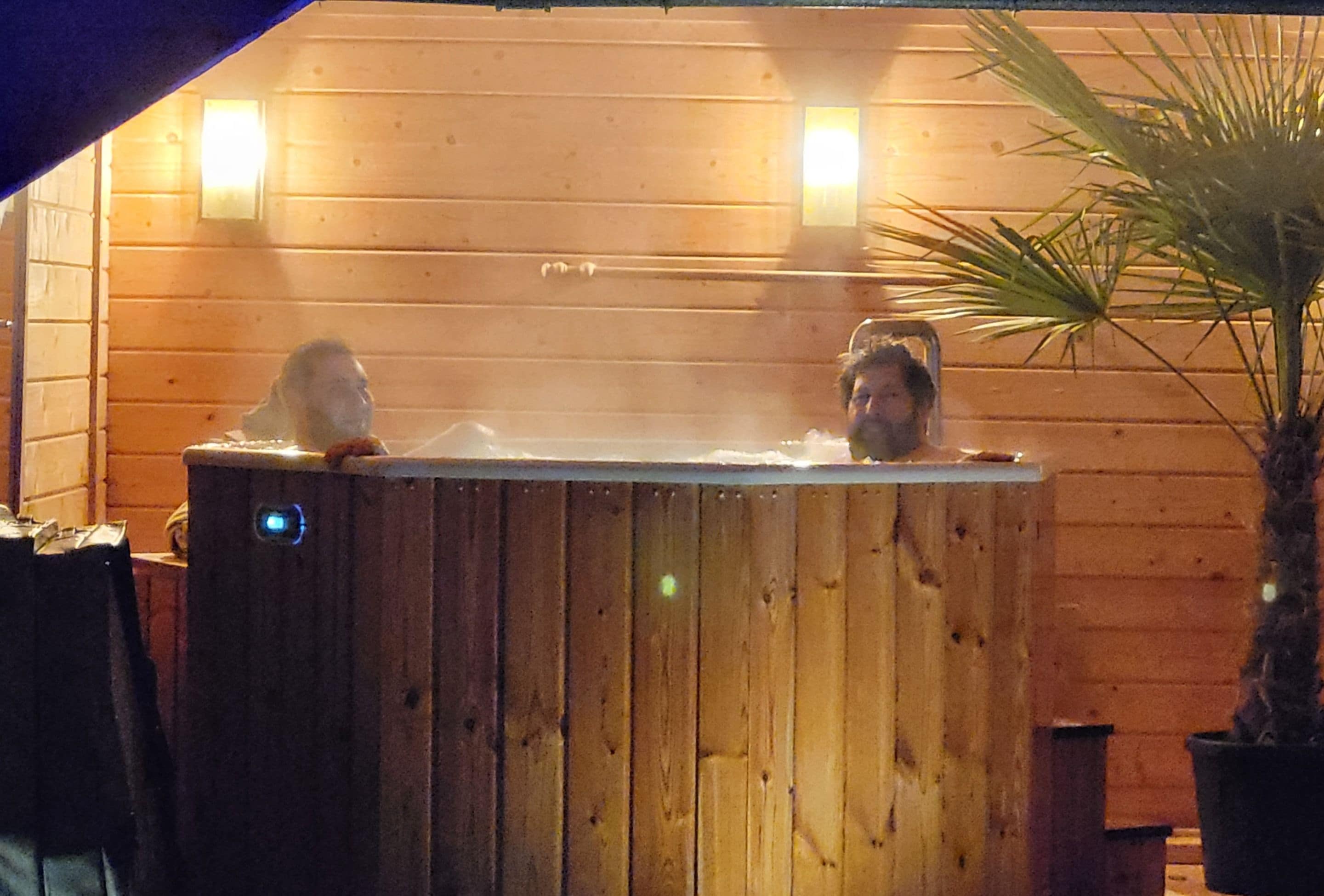 A wooden hot tub with two people inside, surrounded by a wooden wall with two wall-mounted lights.