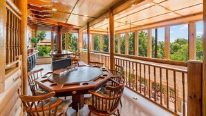 A wooden deck with a round table and chairs, large windows, and a view of greenery.