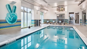 A swimming pool with a blue and white tiled border, a large abstract art piece on the wall, and a seating area with chairs and a table. - Aloft by Marriott Lawrenceville Sugarloaf (Lawrenceville)