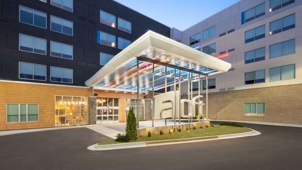 A modern hotel with a glass atrium, a well-lit entrance, and a landscaped area with a small tree. - Aloft by Marriott Lawrenceville Sugarloaf (Lawrenceville)