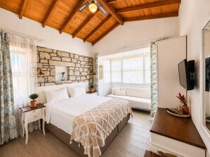A bedroom with a stone wall, wooden ceiling, a bed, bedside tables, a bench, and a TV. - Alacati Fora Bella (Cesme)