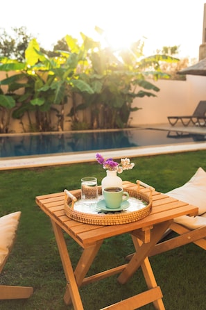 A wooden table with a tray, a cup, and a vase with flowers, set outdoors near a pool.