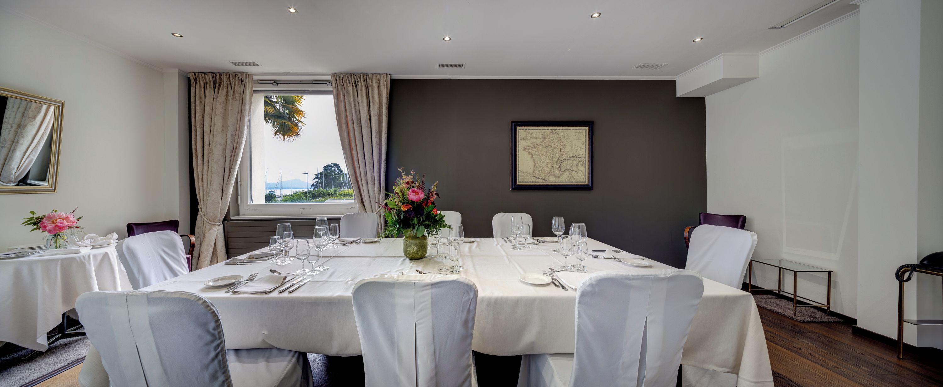 Comedor con una mesa dispuesta para seis, sillas blancas, un florero y un mapa enmarcado en la pared.