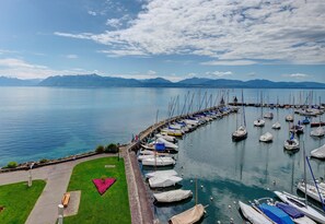 Un puerto deportivo con barcos amarrados a lo largo de un muelle, un área verde con un jardín de flores en forma de corazón, y montañas al fondo.