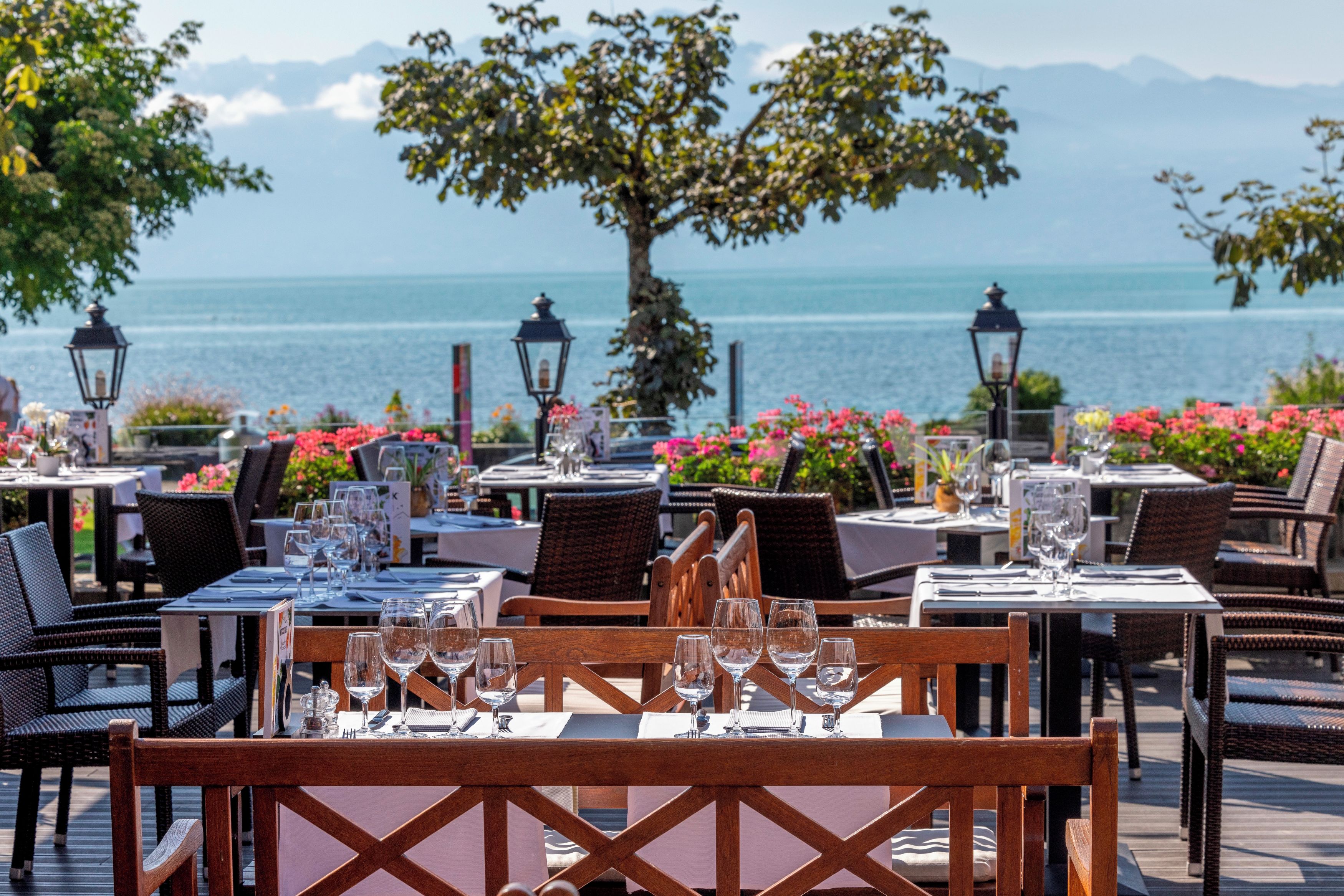Un espace de restauration en plein air, avec des tables dressées pour les clients, offrant une vue sur une étendue d’eau et sur des montagnes au loin.