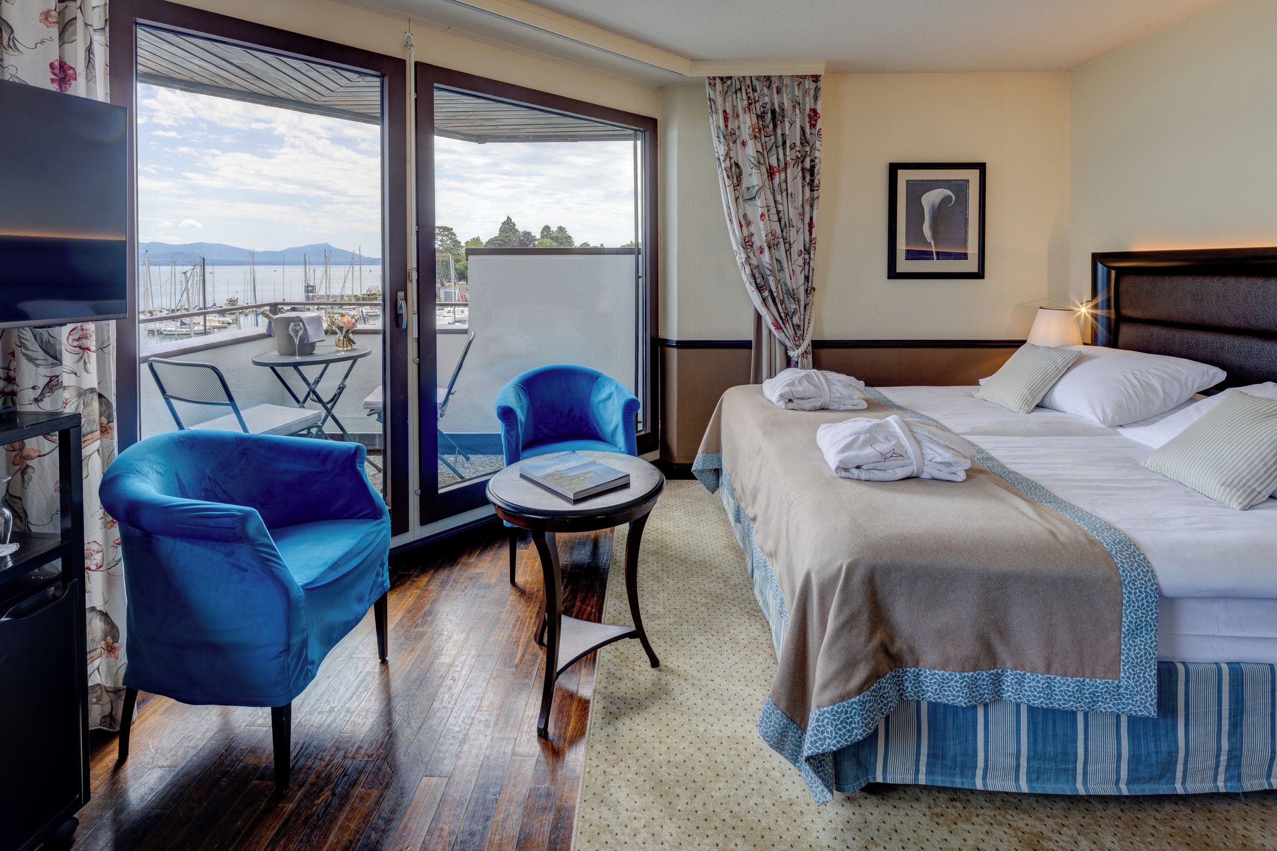 Ein Hotelzimmer mit einem Bett, zwei blauen Sesseln, einem kleinen Tisch, einem Fernseher und Blick auf den Yachthafen.