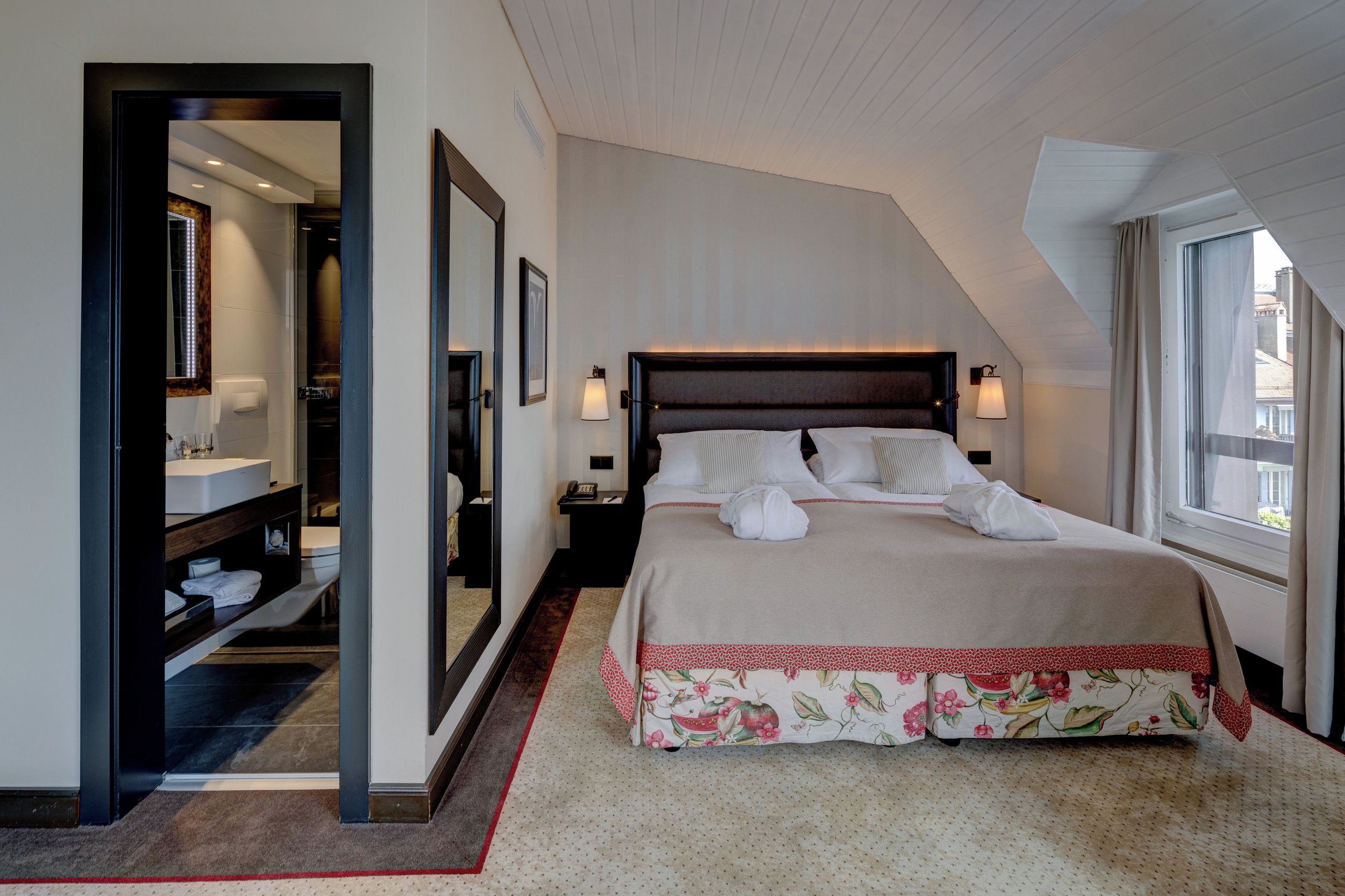 Ein Schlafzimmer mit einem großen Bett, einem Nachttisch, einem Spiegel und Blick nach draußen durch ein Fenster.