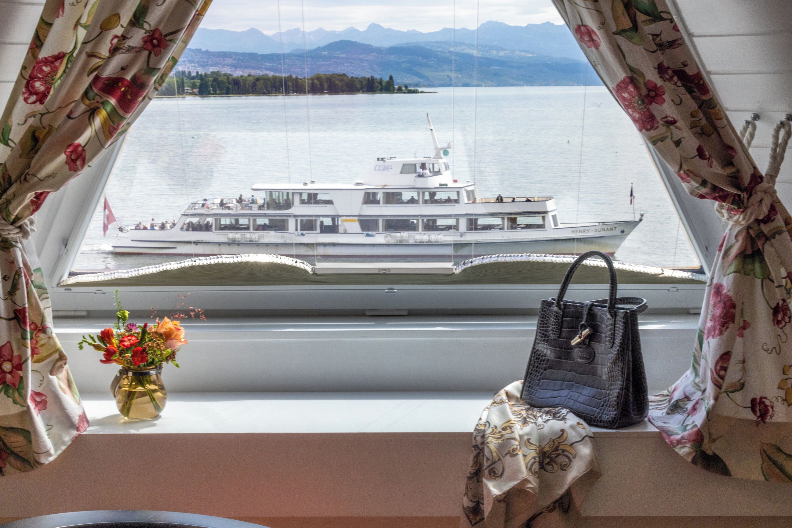 Ein Zimmer mit Seeblick, ein Boot, eine Vase mit Blumen und eine Handtasche auf einem Fensterbrett.