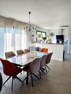 A modern kitchen with a central island, a dining area with a glass-top table, and a chandelier.
