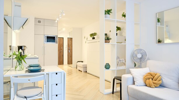 A modern kitchen with a built-in microwave, a coffee machine, and a dining area with a white table and chairs. There is a white sofa with a decorative pillow in the living area. - Apartamenty Sun & Snow Na Wydmach (Dziwnów)