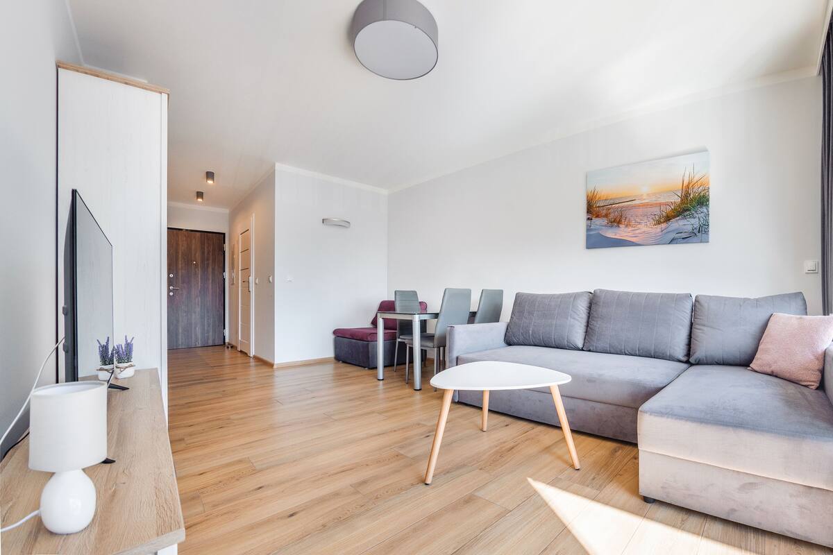 Un salon moderne avec un canapé gris, une petite table blanche et un tableau représentant un coucher de soleil.