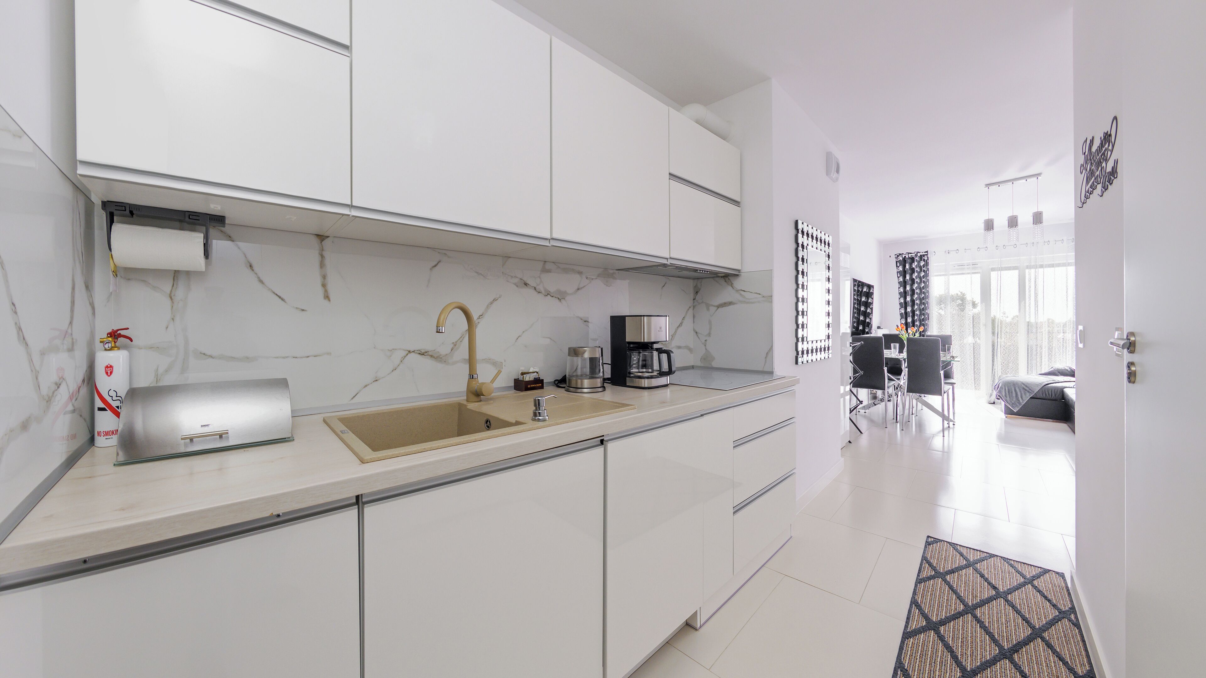 A modern kitchen with white cabinets, a marble backsplash, a sink, a coffee maker, and a dining area with a table and chairs.