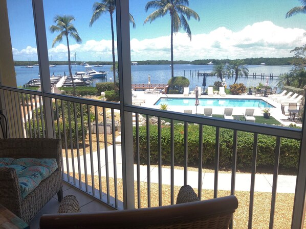 Enjoy the marina view from this balcony with wicker furniture and a railing, overlooking a swimming pool surrounded by lounge chairs and umbrellas.