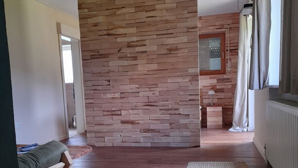 Una habitación con pared de madera, una cama con colchón verde, dos alfombras, un televisor en la pared y una ventana con cortinas.