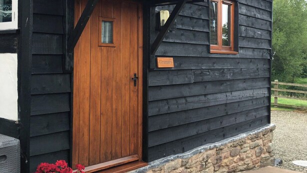 A wooden cabin with a dark exterior, a wooden front door, a small window, and a stone foundation. - 1 bedroom accommodation in Kingsland (Kingsland)