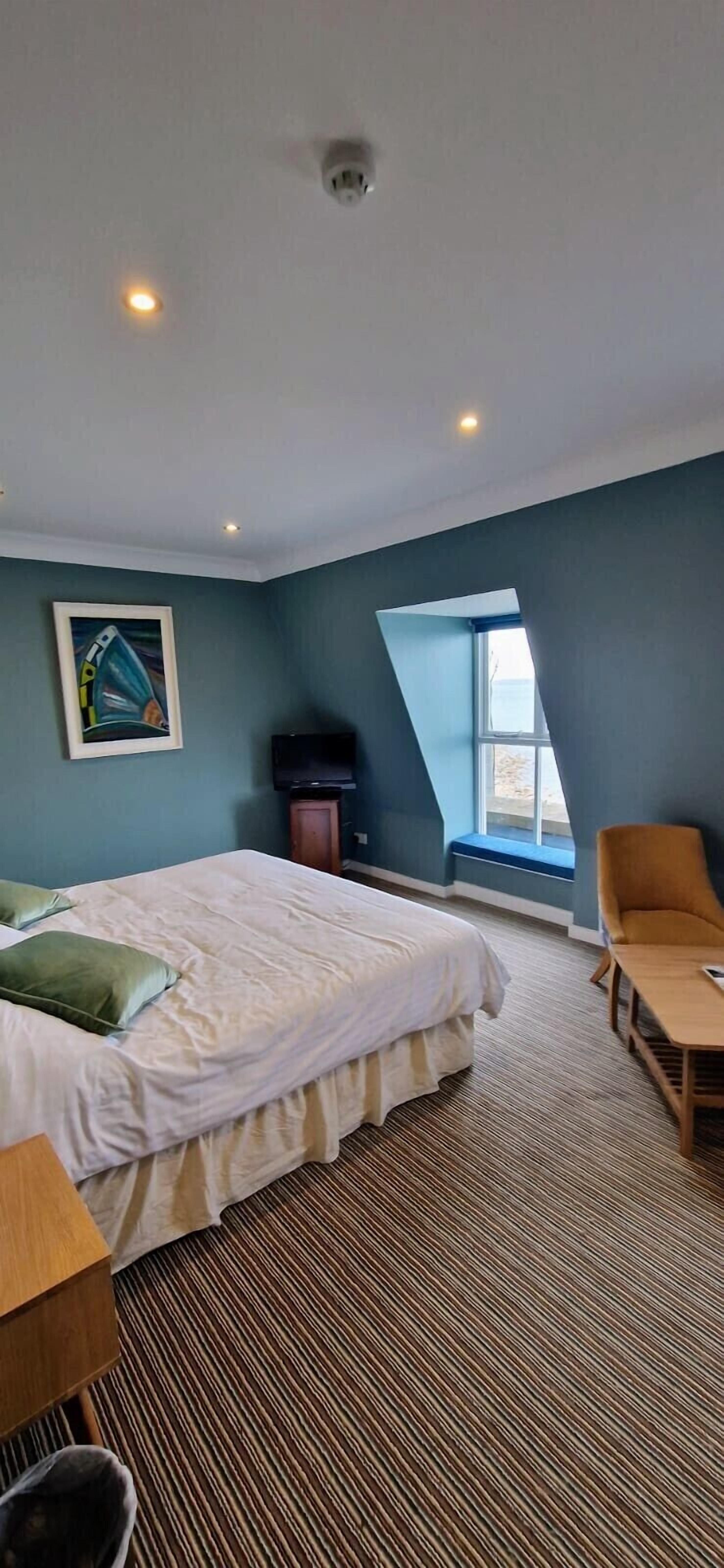 Ett hotellrum med en säng, en stol, ett litet bord, en tv, en tavla, ett fönster och en taklampa.