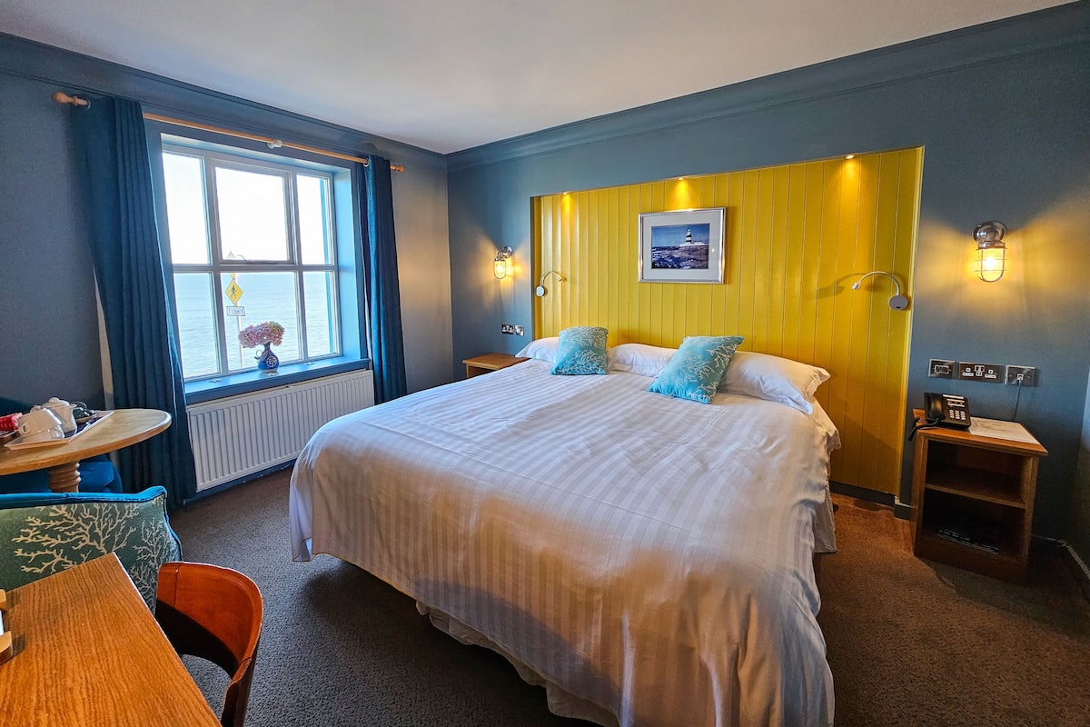 A hotel room with a large bed, a small table, a chair, a window with curtains, and a view of the sea.