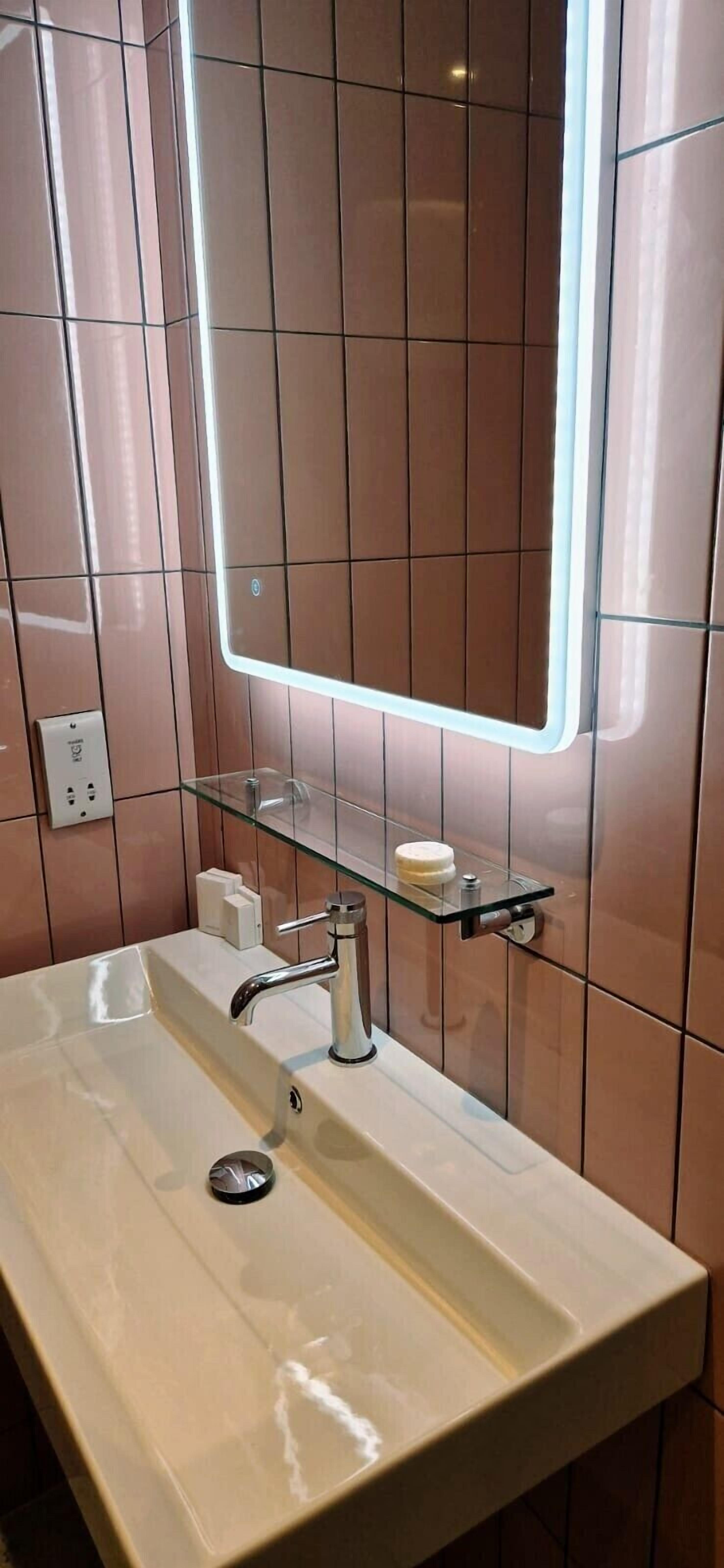 A bathroom with a white sink, a glass shelf with a soap dispenser, and a mirror with a lighted frame.
