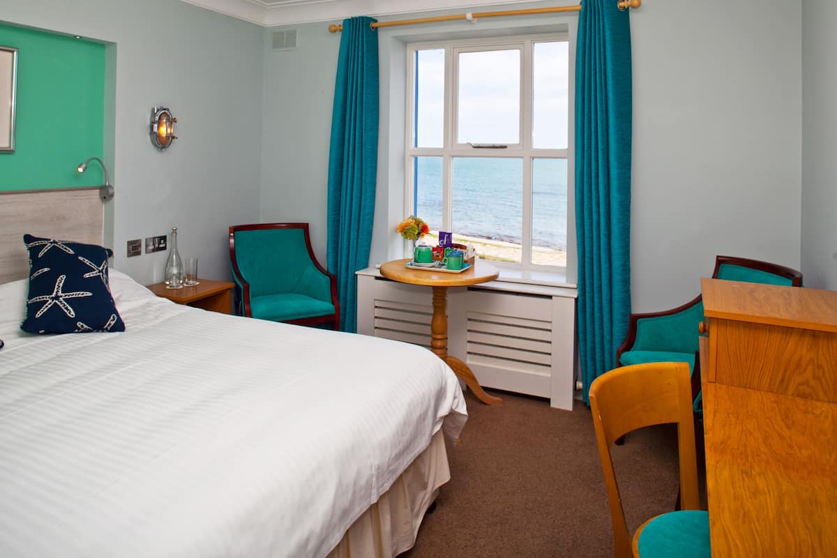 A hotel room with a bed, two chairs, a small table, a window with curtains, a wall-mounted lamp, and a view of the sea.
