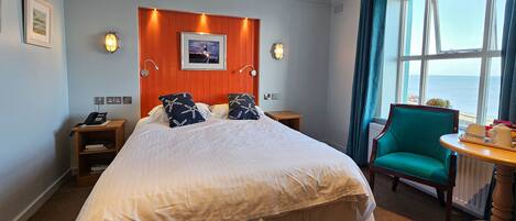 Habitación de hotel con una cama grande, cabecero de madera, dos apliques, una mesita con bandeja, un sillón color turquesa y vista al mar a través de un ventanal amplio.