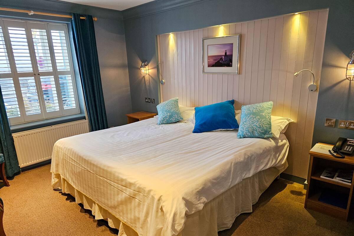 A neatly made bed with a white bedspread and blue pillows, a wooden headboard, a framed picture above the bed, and a bedside table with a lamp and a phone.