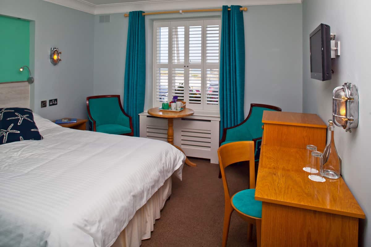 A hotel room with a bed, two chairs, a small table, and a desk with glasses and a bottle.