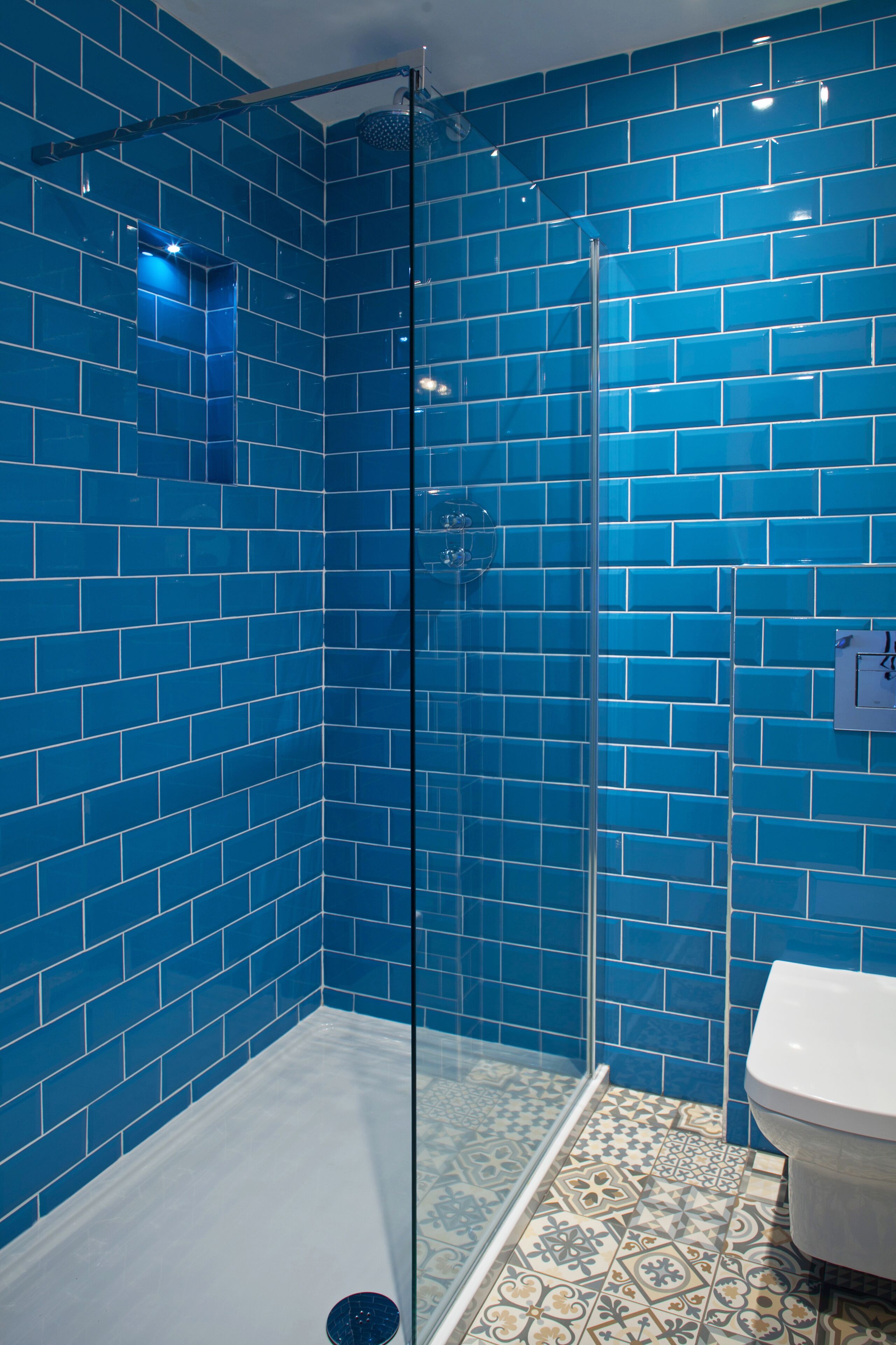 A bathroom with blue tiled walls, a glass shower enclosure, and a toilet with patterned floor tiles.