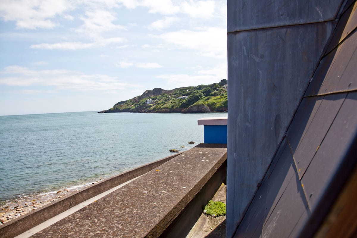 A coastal view with a concrete walkway leading to the sea, a building with a blue facade, and a green hillside in the background.